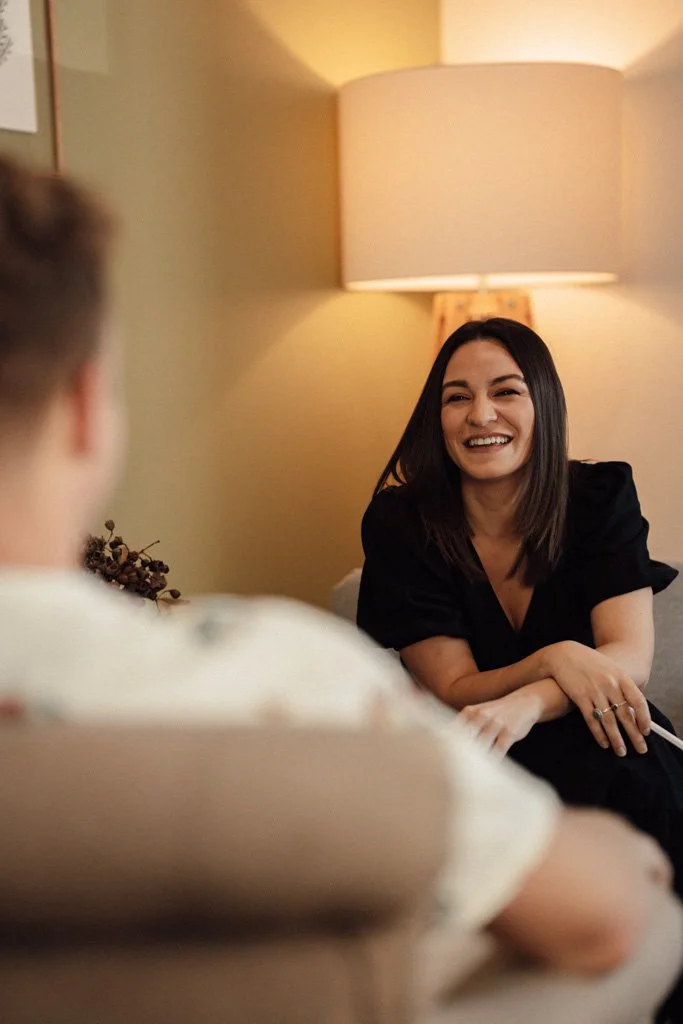 A woman and a man sitting on a couch in a warmly lit room, smiling and talking. Talk therapy, psychology sessions