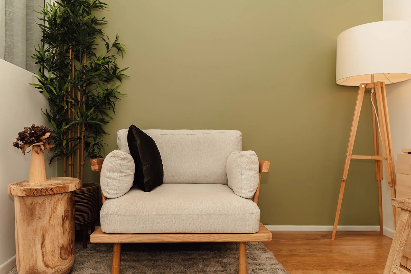Living room with beige loveseat, black and beige pillows, wooden side table with a flower arrangement, green wall, tall potted plant, and wooden floor lamp.